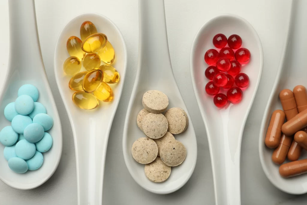 Five white spoons are placed side by side in front of a white background, each holding blue, yellow, brown, and red pills.