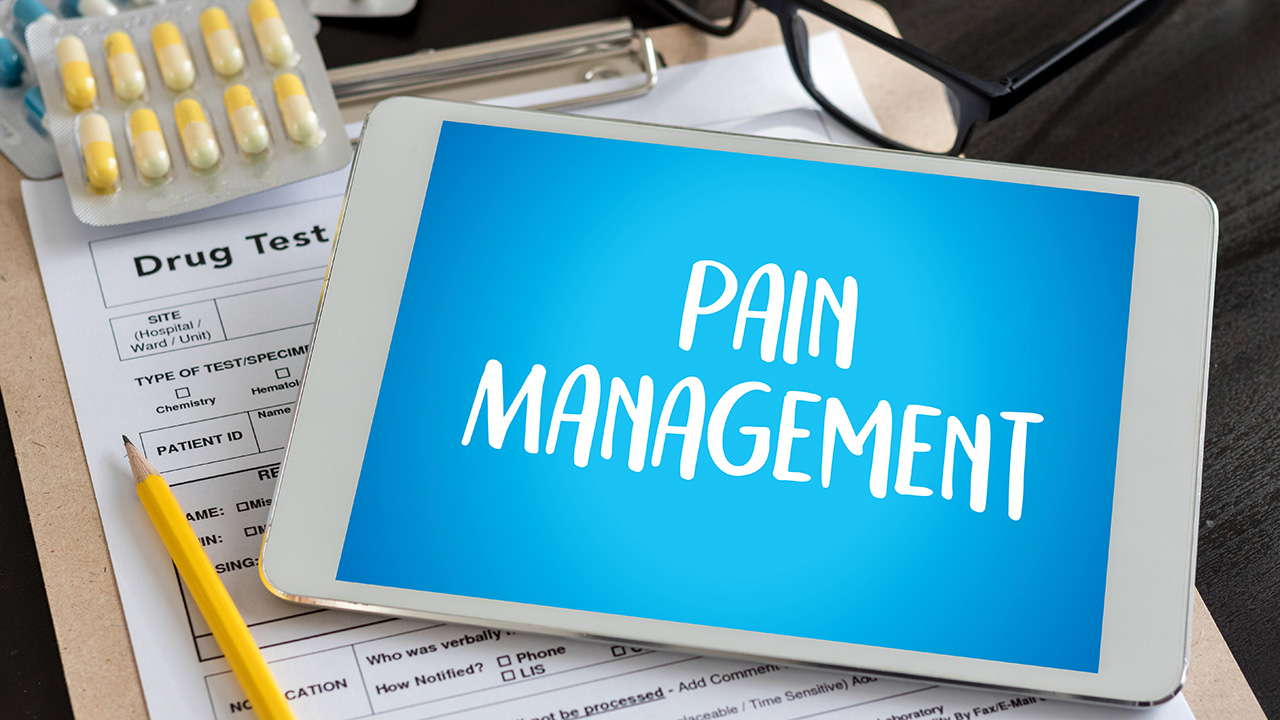 The words “pain management” are displayed in a white font in front of a blue background on an iPad screen, next to a tan clipboard, a yellow pencil, and blue and yellow pills.