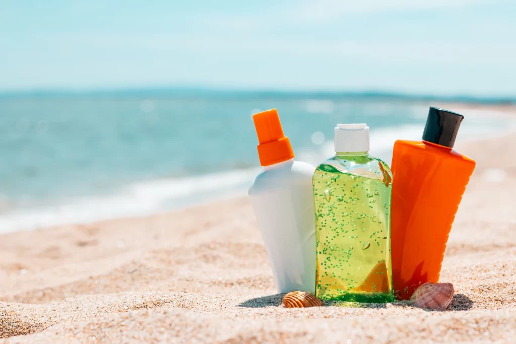 Three bottles of sunscreen spray, aloe vera, and suntan lotion are side-by-side, sitting upright in the sand on the beach next to two small seashells with the ocean in the background.