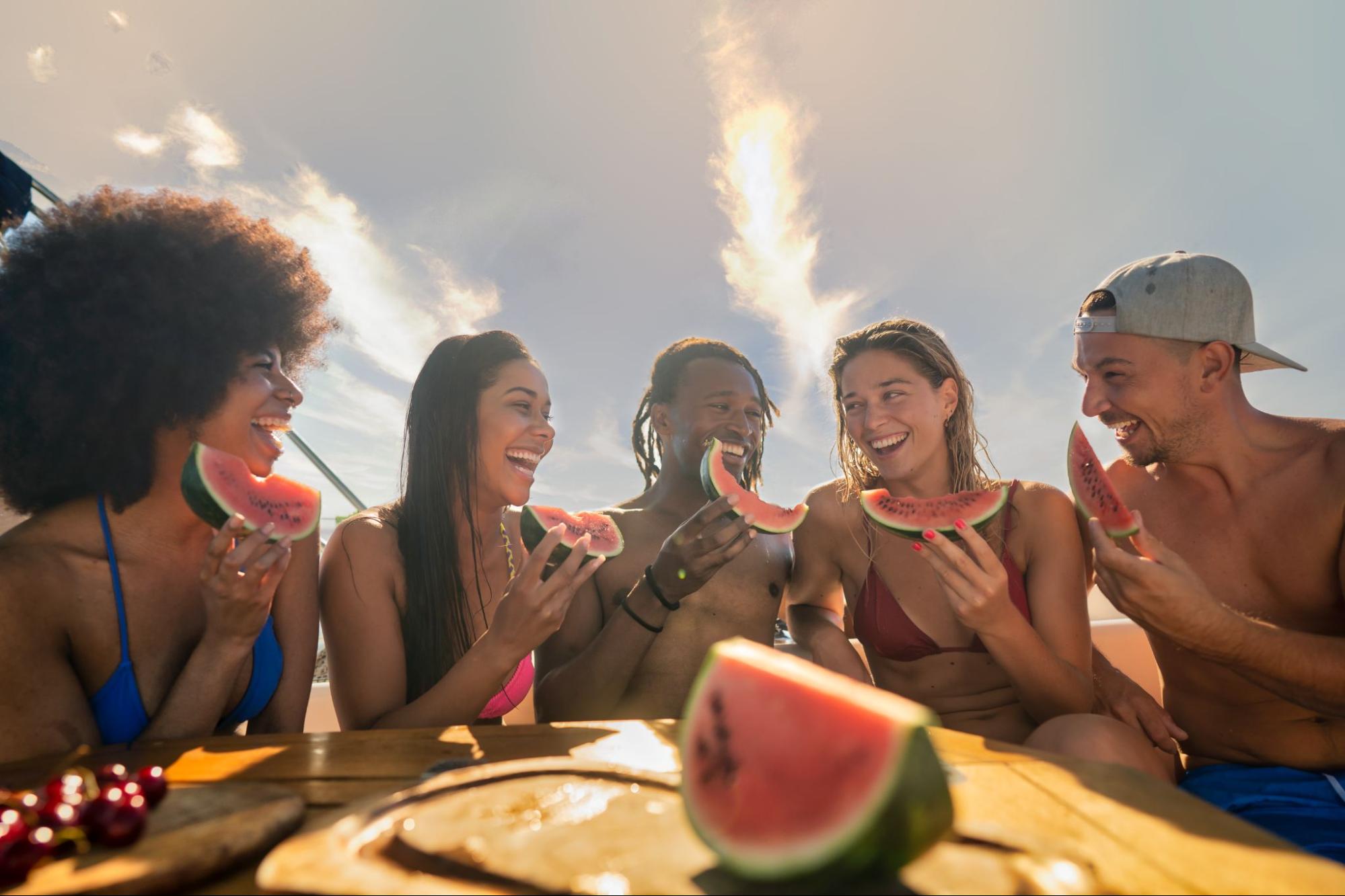 A group of five young adult friends eat watermelon under the hot summer sun.